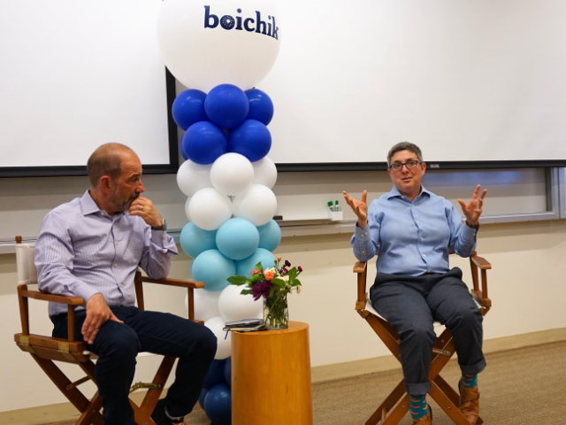 Joe DiNunzio, left, and Emily Winston, right, sit in directors chairs at the front of a classroom, there is a short table with flowers and a balloon tower in between them  