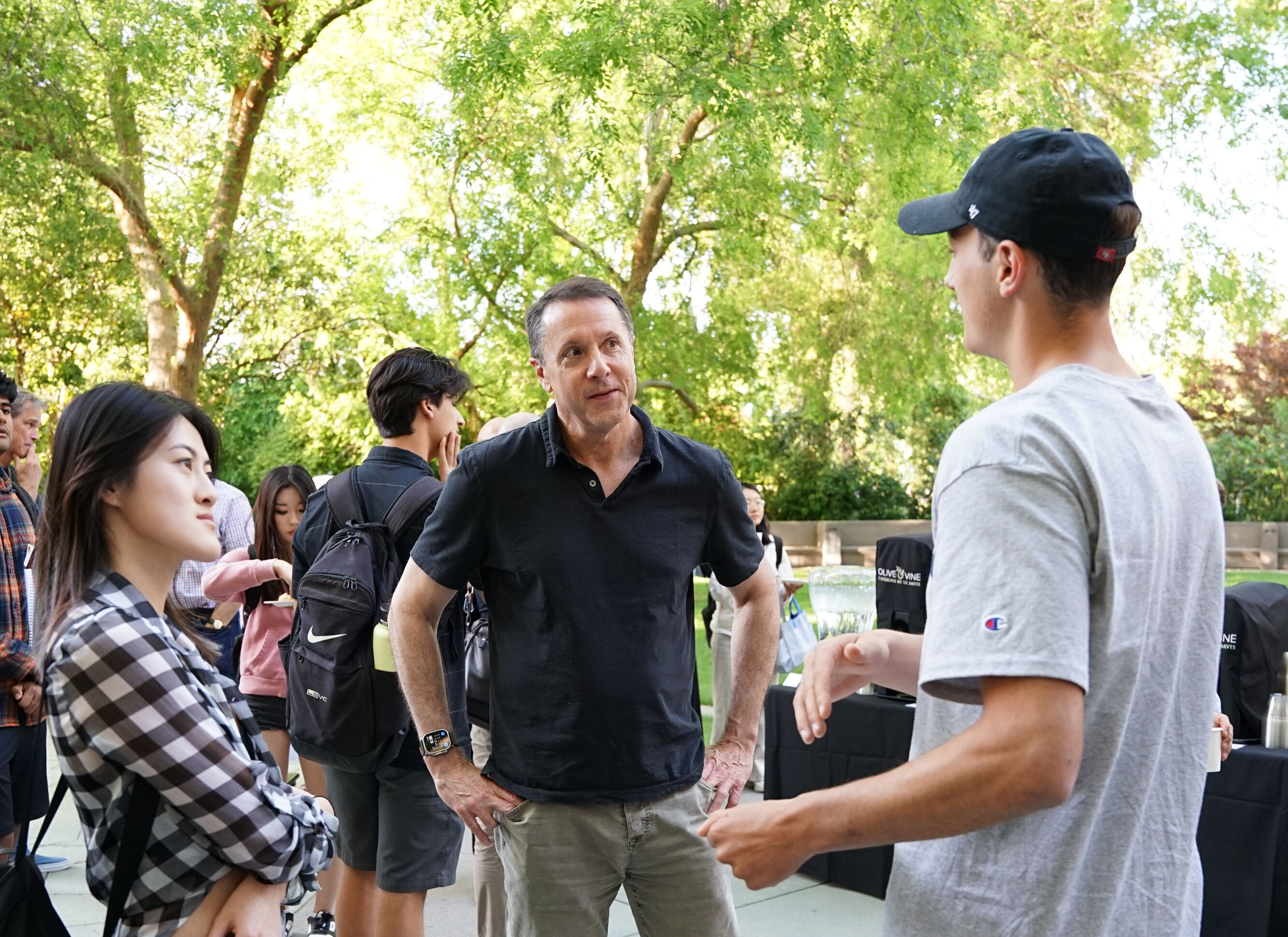 Mitch Laskey stands between two students in an outdoor courtyard. Mitch and the student on the left are looking to the student on the right who appears to be speaking to Mitch.