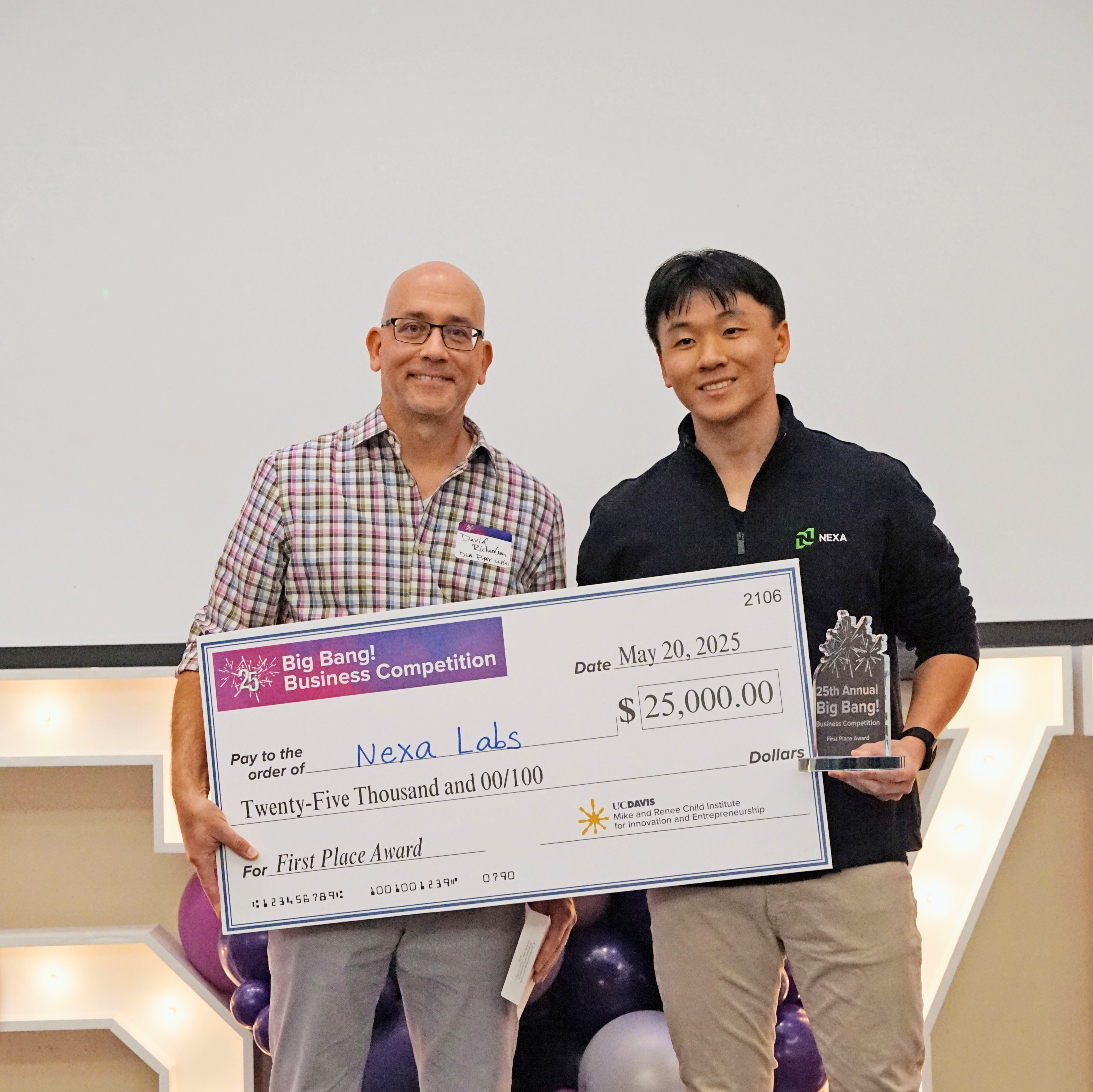 Two males smile while holding a large check made out to Nexa Labs. Male on the right is also holding a Big Bang award
