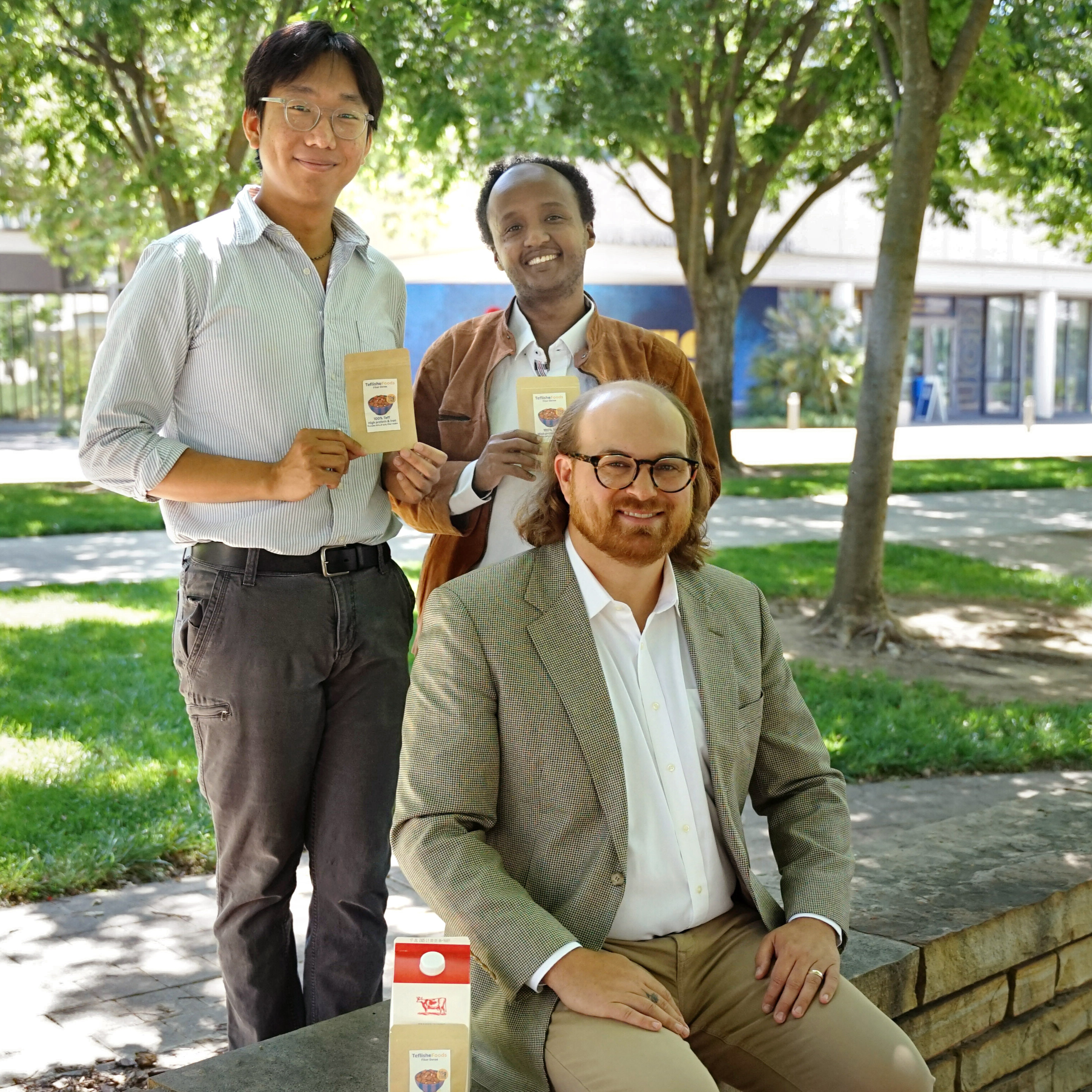 two male students stand behind one male student sitting on stone bench