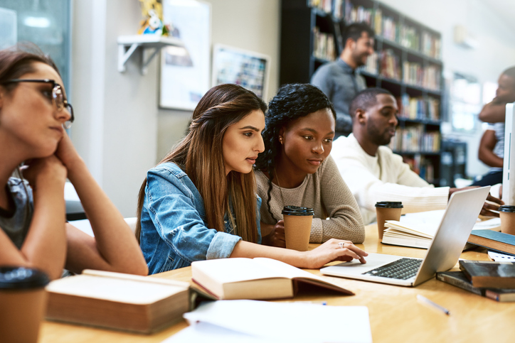 two women students looking at a laptop in a library with other students around them