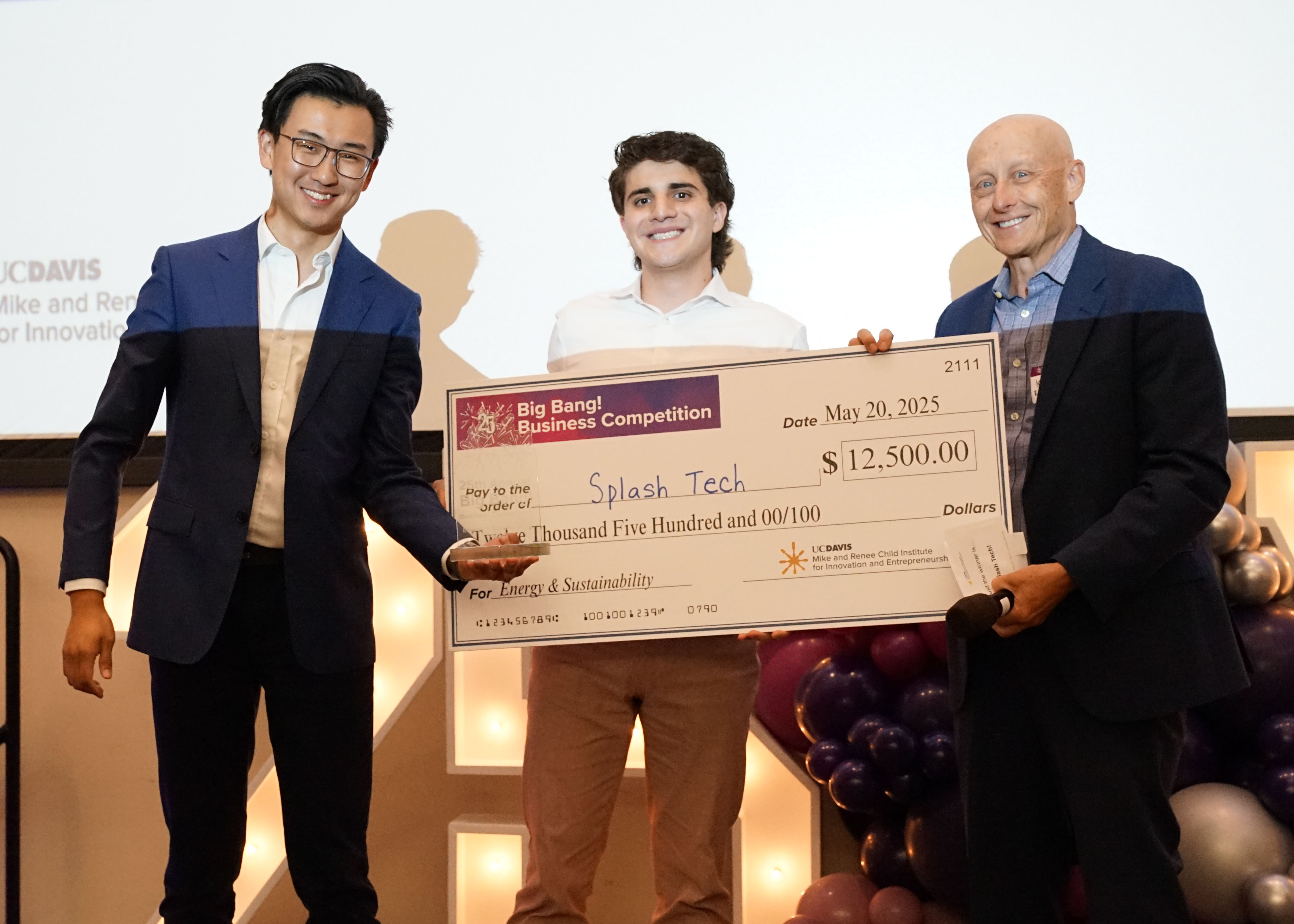 Two students stand on stage, one is holding a large check and the other is holding a physical award in front of the check. A male presenter stands on the right