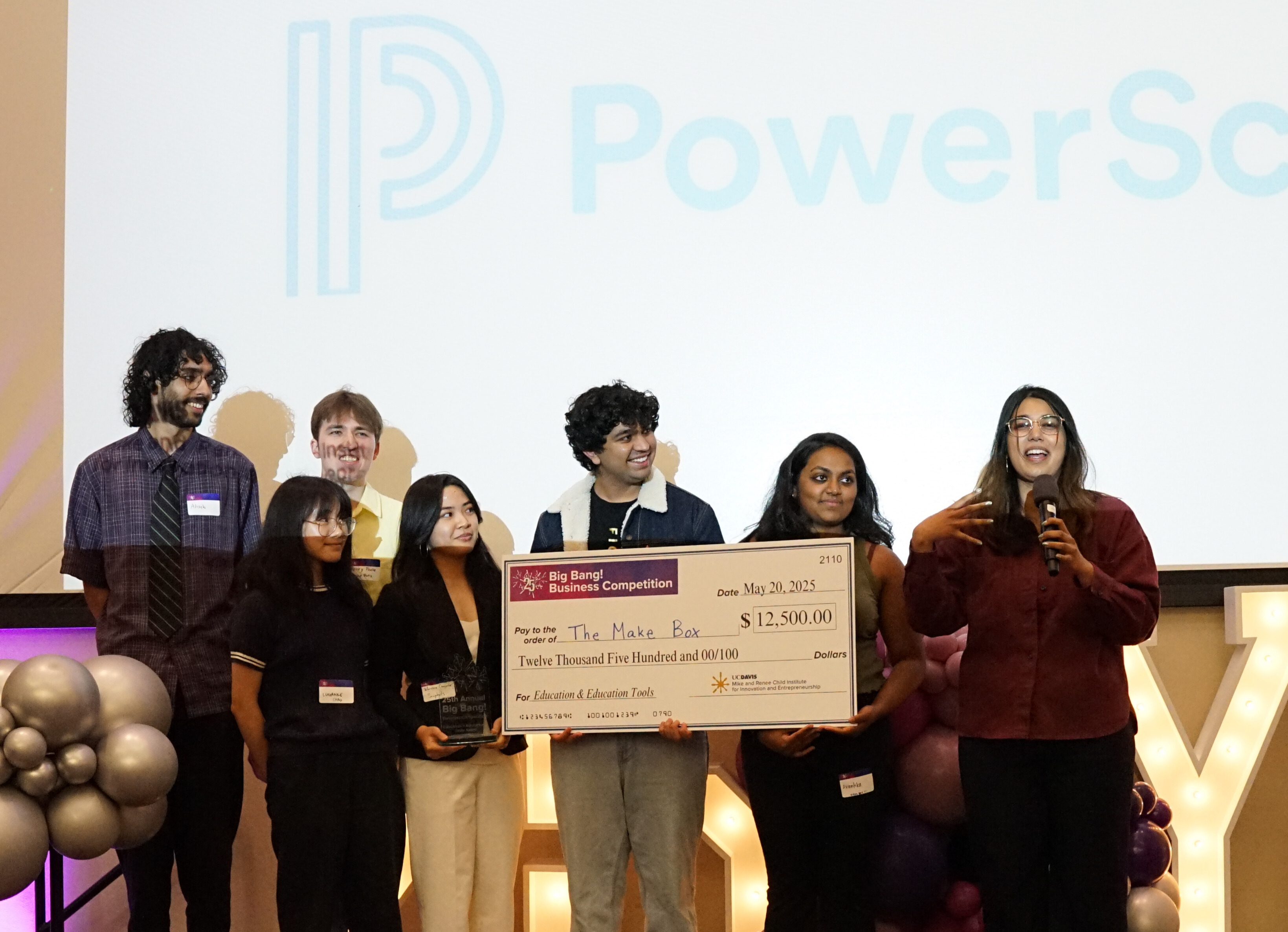 seven students stand on stage to accept their Big Bang award. One student is speaking into a microphone while two other students hold a large check