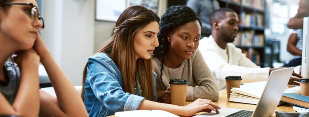 two women students looking at a laptop in a library with other students around them