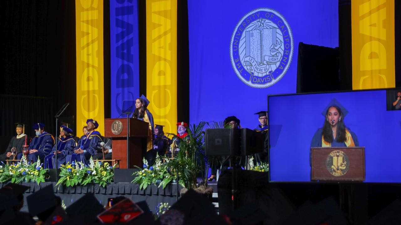 Nishi Baghat speak at a podium at UC Davis commencement. Her image is projected on a large screen to her left.