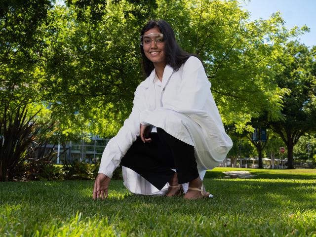 photo of a student wearing a lab coat and PPE crouching down to touch the grass