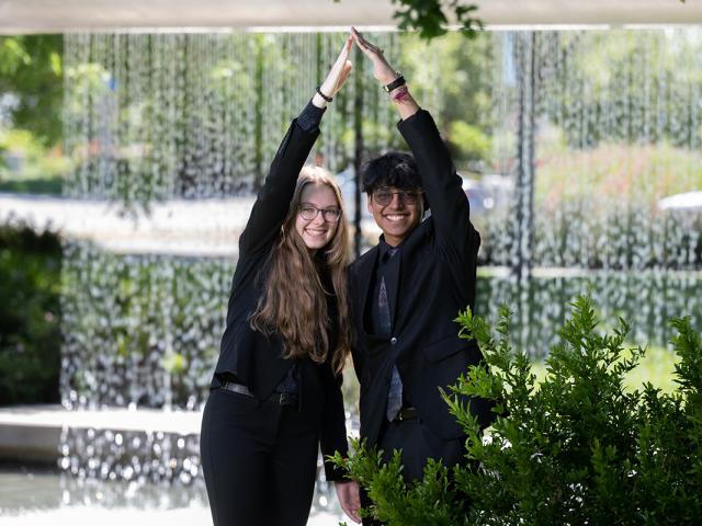 photo of two undergraduates standing in front of a large fountain, forming their logo with their hands and arms