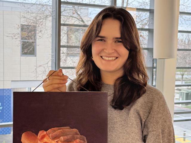 UC Davis EQUIP fellow Juliana smiles and holds up her painting of hands cupping a flame.