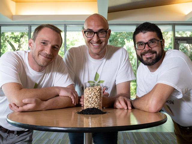 Three men wearing logo t-shirts standing at a table with pistachios and shell by-product