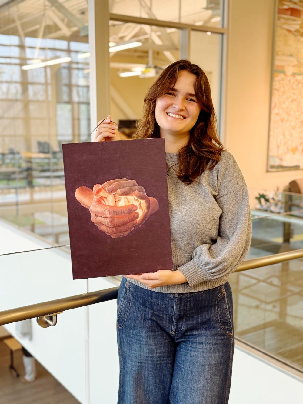 UC Davis EQUIP fellow Juliana smiles and holds up her painting of hands cupping a flame.