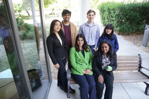 Two students sitting on a bench with four students standing behind them