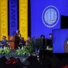 Nishi Baghat speak at a podium at UC Davis commencement. Her image is projected on a large screen to her left.