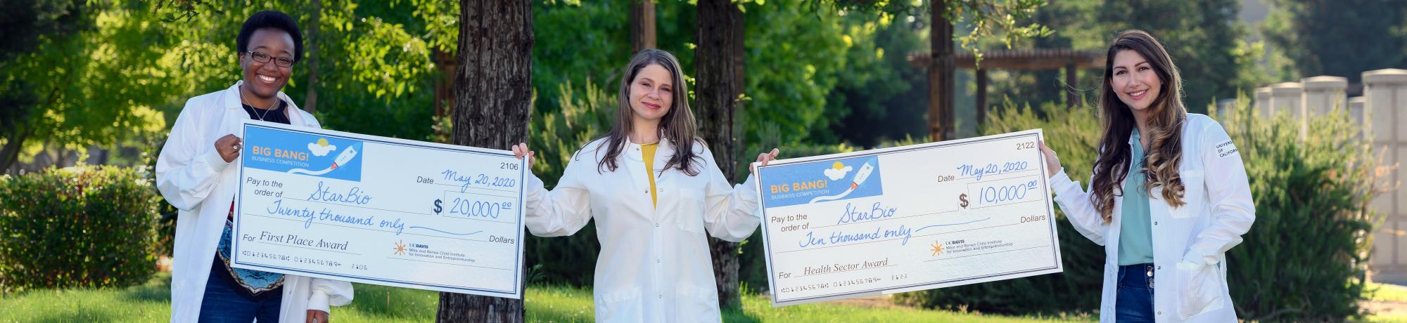 StarBio 2020 Big Bang! winner founders wearing lab coats, smiling while holding large award checks outdoors on grassy hills